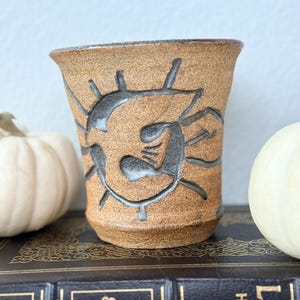 May include: A handmade, brown ceramic tumbler with a textured surface and dark gray abstract design. The cup has a flared top and is displayed on a vintage book. The tumbler is a unique piece of pottery.