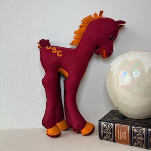 May include: A plush horse toy made of maroon felt with orange accents on the mane, tail, hooves, and muzzle. The horse features the letters USC in orange felt on its hindquarters and has small black bead eyes.