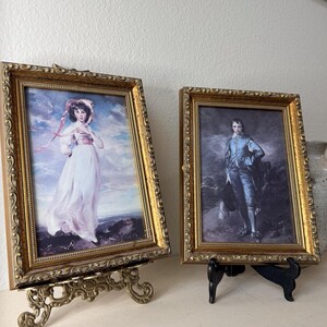 Może przedstawiać: Dwa oprawione portrety w stylu vintage. Jeden przedstawia kobietę w białej sukni i różowej wstążce, drugi mężczyznę w niebieskim garniturze. Oba są w ozdobnych, złotych ramach, eksponowane na dekoracyjnych stojakach.