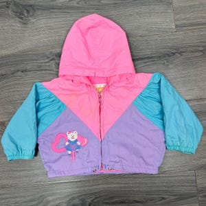 May include: A color-blocked hooded jacket featuring a bright pink upper section, light blue sleeves, and a lavender lower section. A graphic of a white teddy bear wearing a blue dress and riding a bicycle is printed on the lower left side.