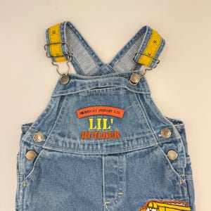 May include: Blue denim overalls with yellow tape measure straps. The overalls feature the text "HIGHWAY REPAIR CO." and "LIL' BUILDER" in orange embroidery. Metal buttons and rivets secure the bib overall design.