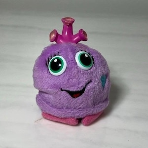 May include: A plush toy in the shape of a purple creature with large, green-eyed eyes and a smiling mouth. It has a pink crown and a small blue heart on its side. The toy is made of soft, fuzzy material.