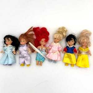 May include: Six small dolls with various hairstyles and outfits are arranged on a white surface. The dolls are dressed in different colors, including blue, purple, pink, yellow, and red. Each doll has a unique facial expression and hairstyle.