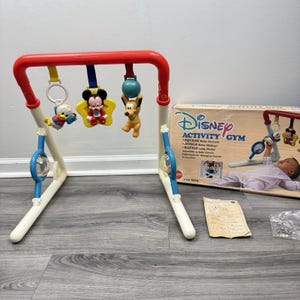 Könnte beinhalten: Ein Vintage Disney Activity Gym für Babys, mit einem rot-weißen Rahmen und hängenden Spielzeugen von Mickey Mouse, Donald Duck und Pluto. Die Box ist sichtbar, mit dem Disney-Logo und Produktdetails.