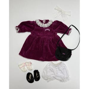 Könnte beinhalten: Ein pflaumenfarbenes Puppenkleid aus Samt mit weißem Spitzenkragen und Manschetten, kombiniert mit einer schwarzen Handtasche, weißen Bloomers, schwarzen Schuhen, weißen Socken mit rosa Schleifen und einer weißen Spitzenschleife. Das Kleid hat ein kleines gesticktes Detail.