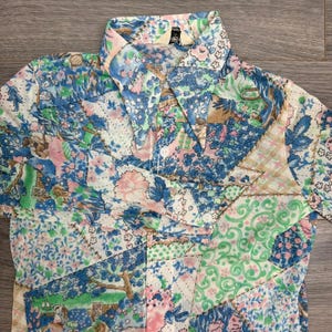 May include: A patchwork shirt featuring a floral design in blue, pink, green, and white. It has a pointed collar and a button-down front. The fabric appears to be a lightweight material.