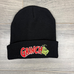 May include: Black knit beanie featuring the word "GRINCH" in red with a green and yellow Grinch character design. The beanie is folded at the brim and is made of a soft, knitted material.