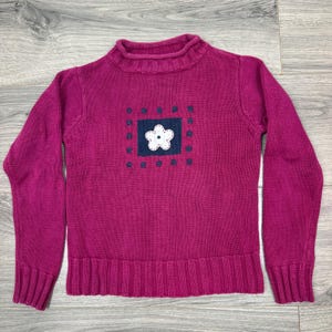Op de afbeelding: Een magenta gebreide trui met lange mouwen, een geribbelde opstaande kraag en een geribbelde zoom. In het midden van de borst zit een donkerblauw rechthoekig embleem met een witte bloemapplicatie, omringd door kleine, donkerblauwe geborduurde bloemetjes.