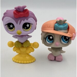 May include: Two colorful owl figurines. One is purple with a red bow and star-shaped eyes, perched on a yellow stand. The other is gray with blue eyes and an orange hat with a blue bow.