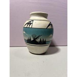 Peut inclure: Petit vase en céramique blanc cassé avec un motif paysager peint. Le vase présente une bande turquoise avec une silhouette noire d'arbres, un tipi et des montagnes. Au-dessus de la bande, des formes géométriques noires.
