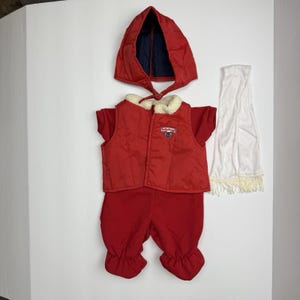 Könnte beinhalten: Ein rotes Baby-Outfit-Set, bestehend aus einer Kapuzenjacke, einer Weste, einer Hose und einem Schal. Die Weste hat einen weißen Kunstfellkragen und ein kleines Emblem. Die Kapuze ist rot mit einem marineblauen Futter. Der Schal ist weiß mit einem Fransenrand.