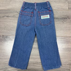 May include: Blue denim pants featuring an elastic waistband, two back pockets with red stitching, and a rectangular white label on the right pocket displaying a green border and a graphic of five small red flowers with green stems and leaves.