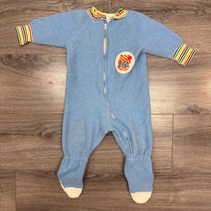 May include: A light blue baby sleeper with a zipper closure, long sleeves, and attached feet. The collar and sleeve cuffs have colorful striped accents. A circular patch with a cartoon design is on the chest.