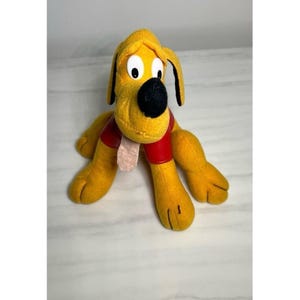 以下が含まれることがあります： 黄色い毛並みの犬のぬいぐるみ。黒い鼻と大きな白い目があります。犬は赤い首輪をしており、ピンクの舌が出ています。おもちゃは座った姿勢で、足を伸ばしています。