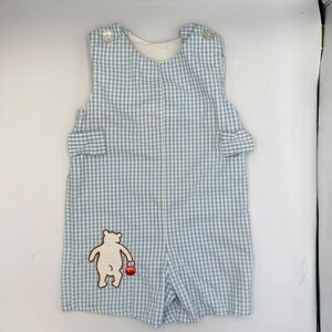 Op de afbeelding: Babyromper in blauw-wit ruitpatroon, met een beerapplicatie met een rode bal. De romper heeft knoopsluitingen op de schouders en een zak. De romper in shorts-stijl is gemaakt van een lichte stof.