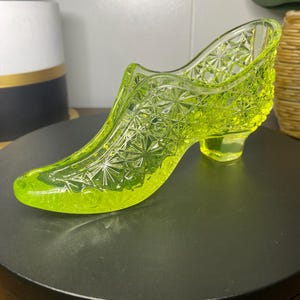May include: A neon green glass slipper with a detailed starburst pattern. The vintage shoe has a small heel and a pointed toe. The glass is transparent, allowing light to pass through, and is displayed on a black surface.