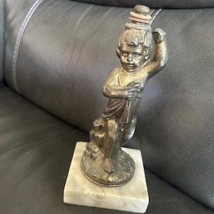 May include: A bronze-toned statue of a cherubic figure holding a cloth, mounted on a white marble base. The statue has a detailed design and is approximately 6 inches tall. The figure is wearing a crown.