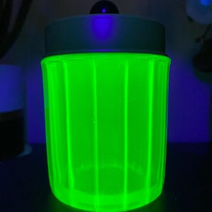 May include: A glowing green glass jar with a light blue lid and a small black knob on top. The jar has vertical ridges and is illuminated with a bright green light, creating a striking visual effect. The jar is cylindrical in shape.
