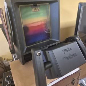 Könnte beinhalten: Eine schwarze Vectrex-Spielkonsole mit einer Star Wars-Bildschirmauflage und einem schwarzen Steuerjoystick, auf dessen Vorderseite der Text Yoke Vectrex aufgedruckt ist. Die Konsole hat einen vertikalen Monitor und ein rechteckiges Gehäuse.