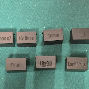 Puede incluir: Un conjunto de siete adaptadores rectangulares de plástico negro con texto grabado. Las etiquetas incluyen Minox x2, 16+8mm, 16mm, 17.5, 21mm, Eljy 30 y Kombi 31.6. Estos pequeños componentes están diseñados para equipos de cine o cámaras.