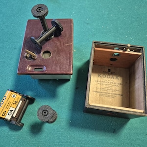 Può includere: Un corpo macchina fotografica vintage Pocket Kodak con rivestimento testurizzato marrone scuro, un telaio interno in legno separato con testo stampato, due bobine di pellicola in plastica nera e una cartuccia di pellicola 35mm gialla GA 36 exp.