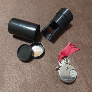 May include: A vintage black and silver film camera with a red ribbon attached. The camera is in a black case with a black lid. The camera has a silver lens and a silver dial with the word "PETAU" on it.