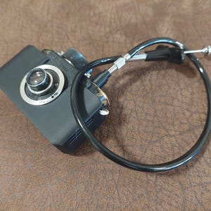 May include: A black vintage camera with a silver lens and a black cable attached to it.