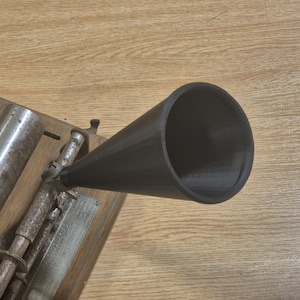以下が含まれることがあります： 黒いホーンスピーカーを備えたヴィンテージ蓄音機。蓄音機は金属部品を備えた木製のベースに取り付けられています。ホーンは円錐形で右を向いています。木製のベースは、明るい茶色の木製の表面にあります。