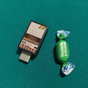 Puede incluir: Una pequeña radio retro negra y marrón con un diseño de arcoíris y una memoria USB conectada. Junto a ella hay un caramelo envuelto en verde y blanco con el texto "Eucalipto & Mentol" en él.