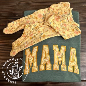 Op de afbeelding: Een groen sweatshirt met het woord "MAMA" geborduurd in geel met een bloemenpatroon. Een bijpassende, bloemige baby-romper ligt op het sweatshirt.