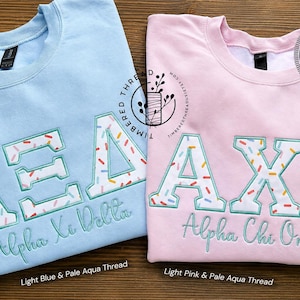 May include: A light blue sweatshirt with embroidered Alpha Xi Delta Greek letters and a light pink sweatshirt with embroidered Alpha Chi Omega Greek letters. Both feature white letters with colorful sprinkles and pale aqua thread outlines.
