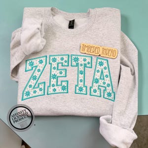 May include: A light gray sweatshirt with the word "ZETA" embroidered in teal with white floral accents. The sweatshirt has a crew neck and long sleeves. A wooden tag reads "TIMBERED THREAD". An "Affinity Licensed Product" sticker is visible.