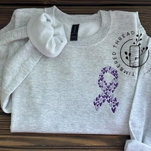 May include: A light grey crewneck sweatshirt featuring an embroidered awareness ribbon on the left chest, composed of small purple and lavender hearts. A matching light grey fabric scrunchie rests on the collar area of the garment.