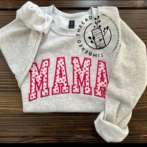 May include: Gray sweatshirt with the word "MAMA" in red letters with a pink and white heart pattern. The sweatshirt has a small logo on the sleeve that says "Timbered Thread".