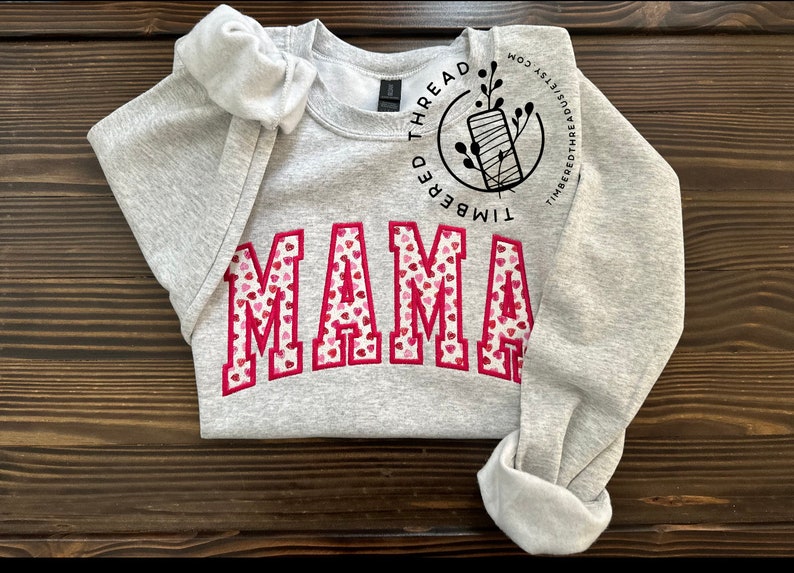 May include: Gray sweatshirt with the word "MAMA" in red letters with a pink and white heart pattern. The sweatshirt has a small logo on the sleeve that says "Timbered Thread".