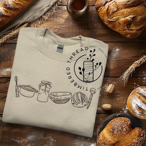 Op de afbeelding: Een beige sweatshirt met een zwart geborduurd ontwerp van keukengerei en de tekst "TIMBERED THREAD" in een cirkel. De tekst "WOOD AS TIMBERED THREAD.COM" is ook geborduurd op het sweatshirt.