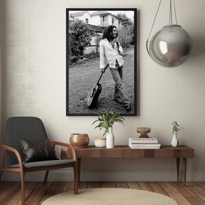 Op de afbeelding: Zwart-witfoto van een man met dreadlocks die een gitaar vasthoudt. Hij draagt een wit shirt en een spijkerbroek. De foto is ingelijst in een zwart frame.