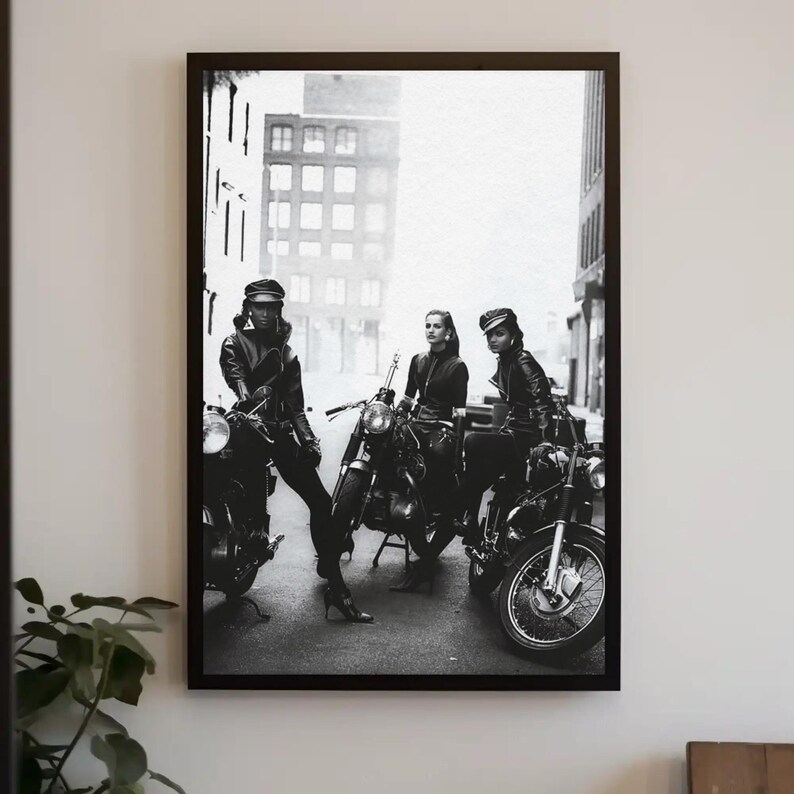 May include: Black and white framed print featuring three people posing with motorcycles. They are wearing leather jackets, caps, and boots. The motorcycles are parked on a city street, with buildings in the background.