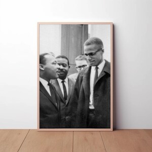 May include: A framed black and white photograph showing four men in suits. The central figure, wearing glasses and a dark coat, is surrounded by others in suits and ties. The image is set against a neutral background, with a wooden floor.