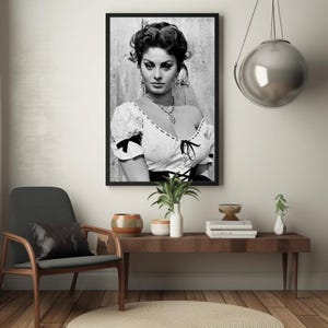 May include: Black and white portrait photograph of a woman with dark hair and a white blouse. The woman is looking directly at the camera. The photograph is framed in a black frame.