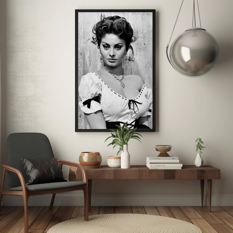 May include: Black and white portrait photograph of a woman with dark hair and a white blouse. The woman is looking directly at the camera. The photograph is framed in a black frame.
