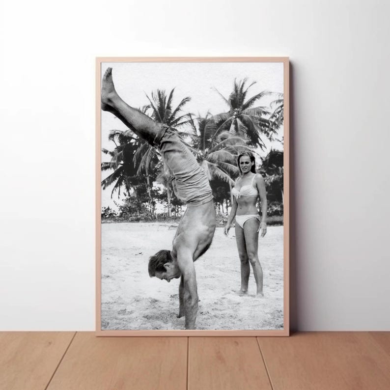 Op de afbeelding: Een zwart-witfoto van een man die handstand maakt op een strand, met een vrouw die achter hem staat. De man draagt ​​jeans en de vrouw een bikini. Palmbomen zijn zichtbaar op de achtergrond.