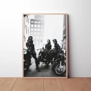 May include: Black and white framed print featuring three people posing with motorcycles. The people are wearing leather jackets and hats. The motorcycles are parked on a city street. The print is in a wooden frame.