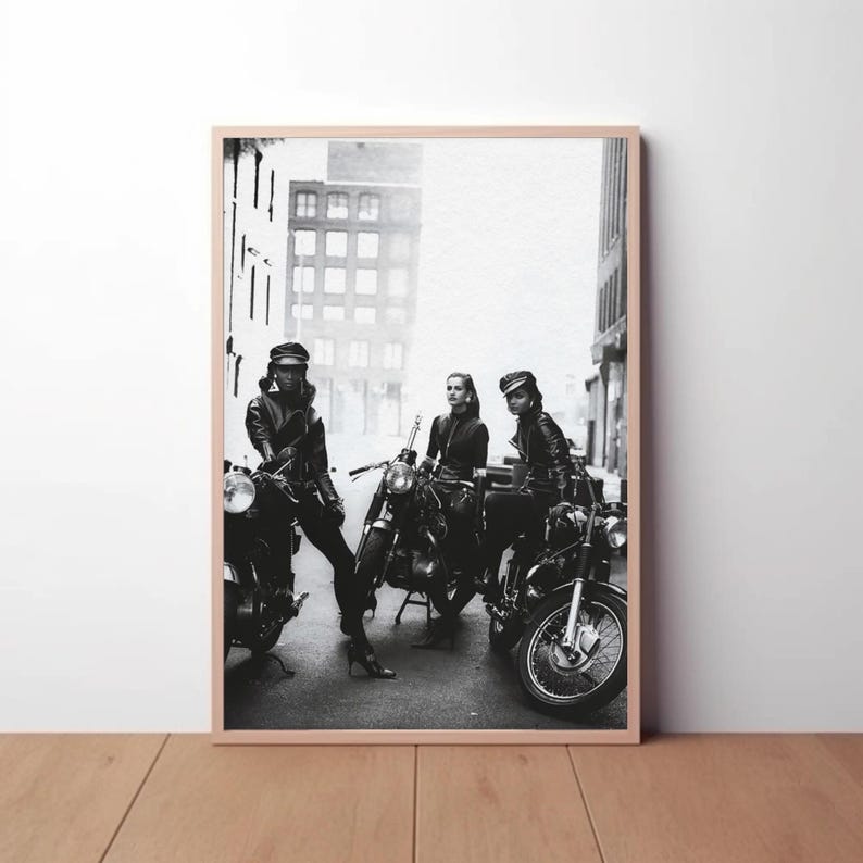 May include: Black and white framed print featuring three people posing with motorcycles. The people are wearing leather jackets and hats. The motorcycles are parked on a city street. The print is in a wooden frame.