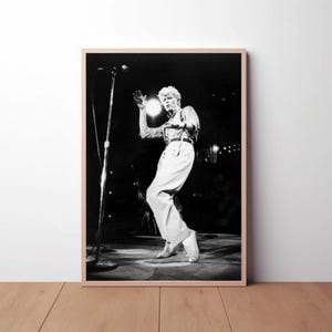 Peut inclure: Photographie en noir et blanc d'un musicien se produisant sur scène. Le musicien porte une chemise et un pantalon blancs et tient un microphone. La scène est sombre et le musicien est éclairé par un projecteur.