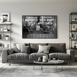 May include: A monochrome framed print depicting an Italian restaurant with the sign reading "Italian Kitchen Spaghetti 30&cent; Beer-Wine Pizzeria". A grey sofa with cushions, a round coffee table, and a rug are in the foreground. Shelving units with decor are on either side.