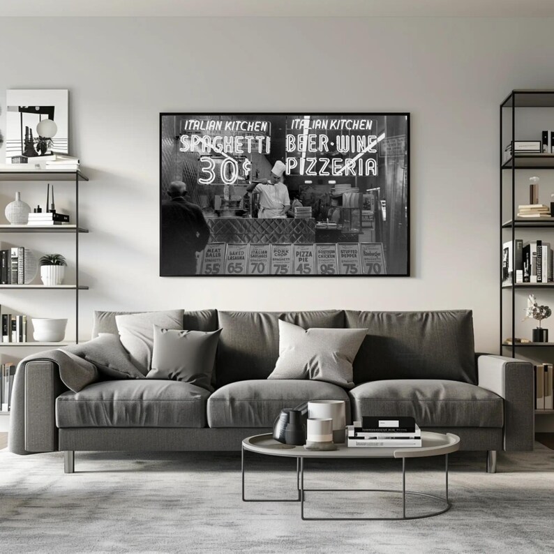 May include: A monochrome framed print depicting an Italian restaurant with the sign reading "Italian Kitchen Spaghetti 30&cent; Beer-Wine Pizzeria". A grey sofa with cushions, a round coffee table, and a rug are in the foreground. Shelving units with decor are on either side.