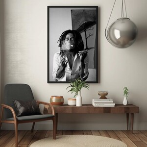 Pu&ograve; includere: Fotografia in bianco e nero di un uomo con i capelli lunghi che fuma una sigaretta. La foto &egrave; incorniciata da una cornice nera e appesa a un muro bianco. C'&egrave; un tavolo di legno con un vaso di piante e un libro sopra.