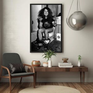 May include: Black and white photo of a man sitting on a toilet, holding a glass of liquid and a cigarette. The photo is framed in a black frame and hanging on a white wall.