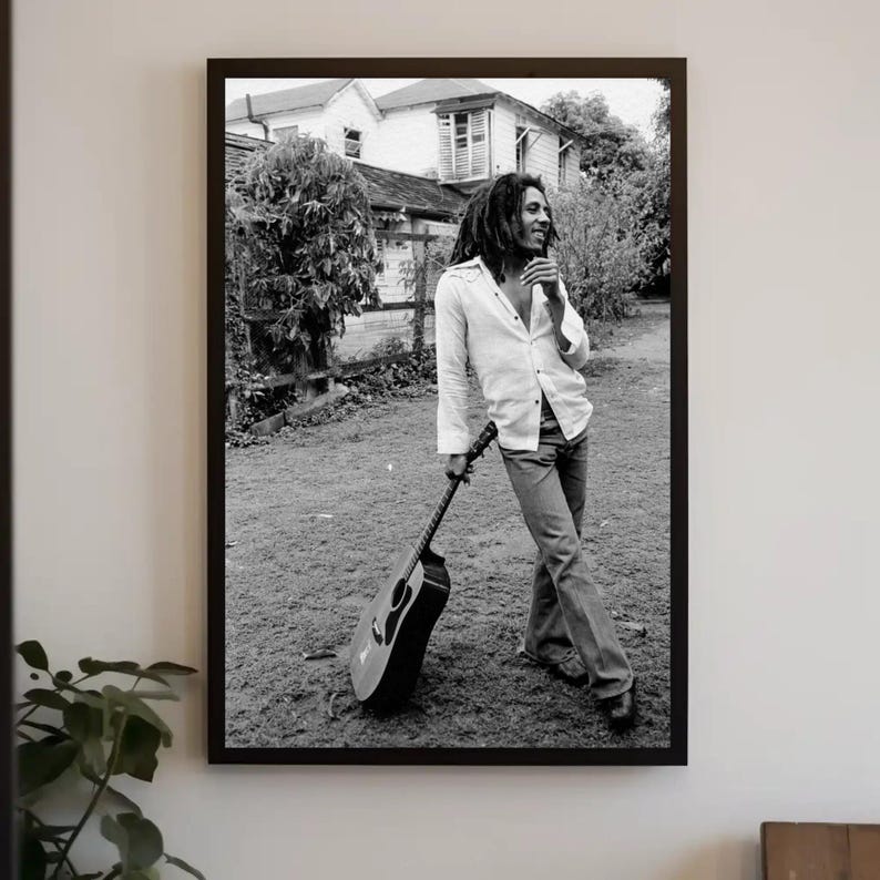 Op de afbeelding: Zwart-witfoto van een man met lang haar en een baard die een gitaar vasthoudt. Hij draagt een wit overhemd en een spijkerbroek. De foto is gemaakt buiten voor een huis.